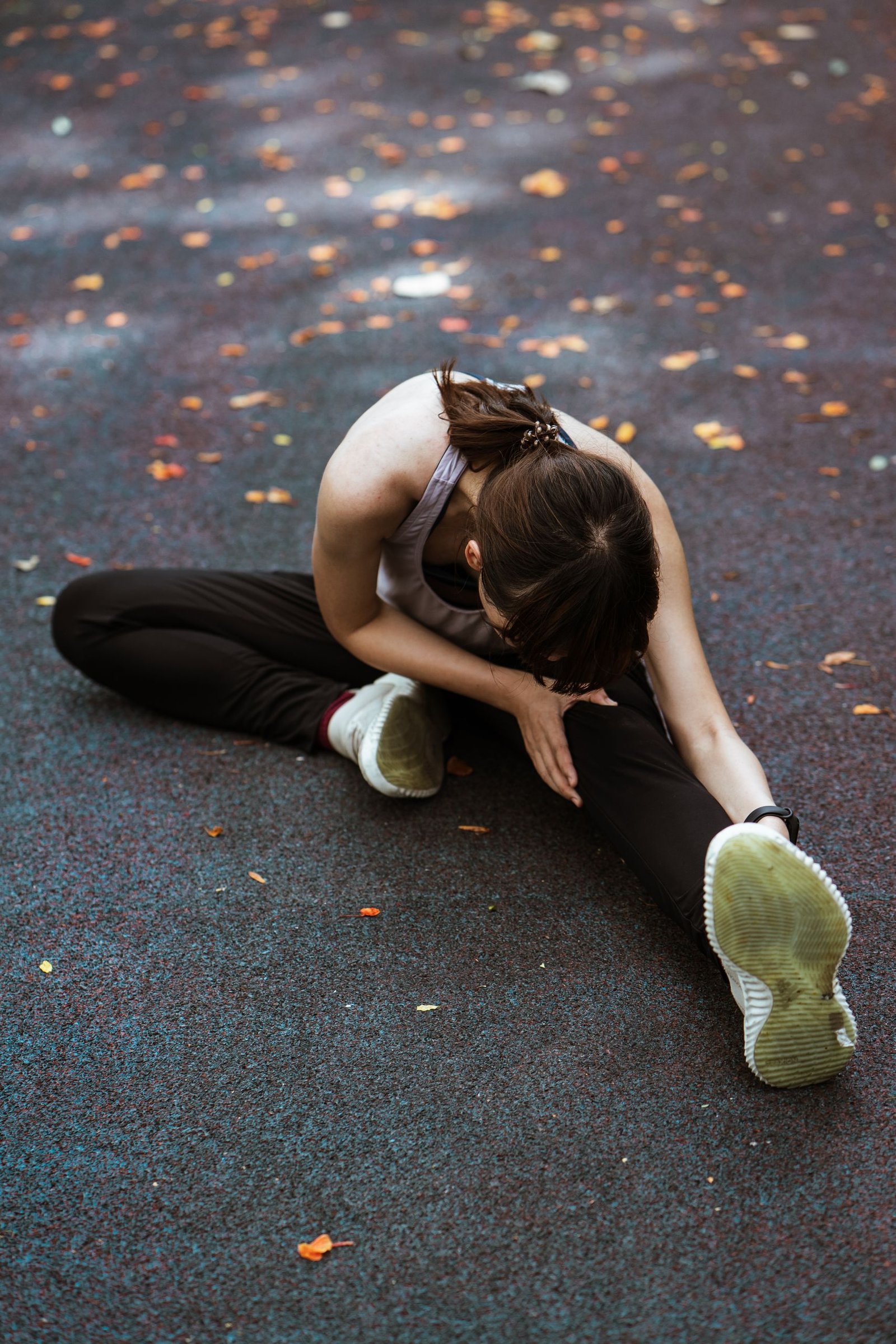 Anonymous flexible female in sportswear and sneakers sitting on pavement and bending forward while stretching on sports ground